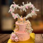 Elegant pink birthday cake with floral decorations, feathers, and a crown topper, perfect for celebrations.